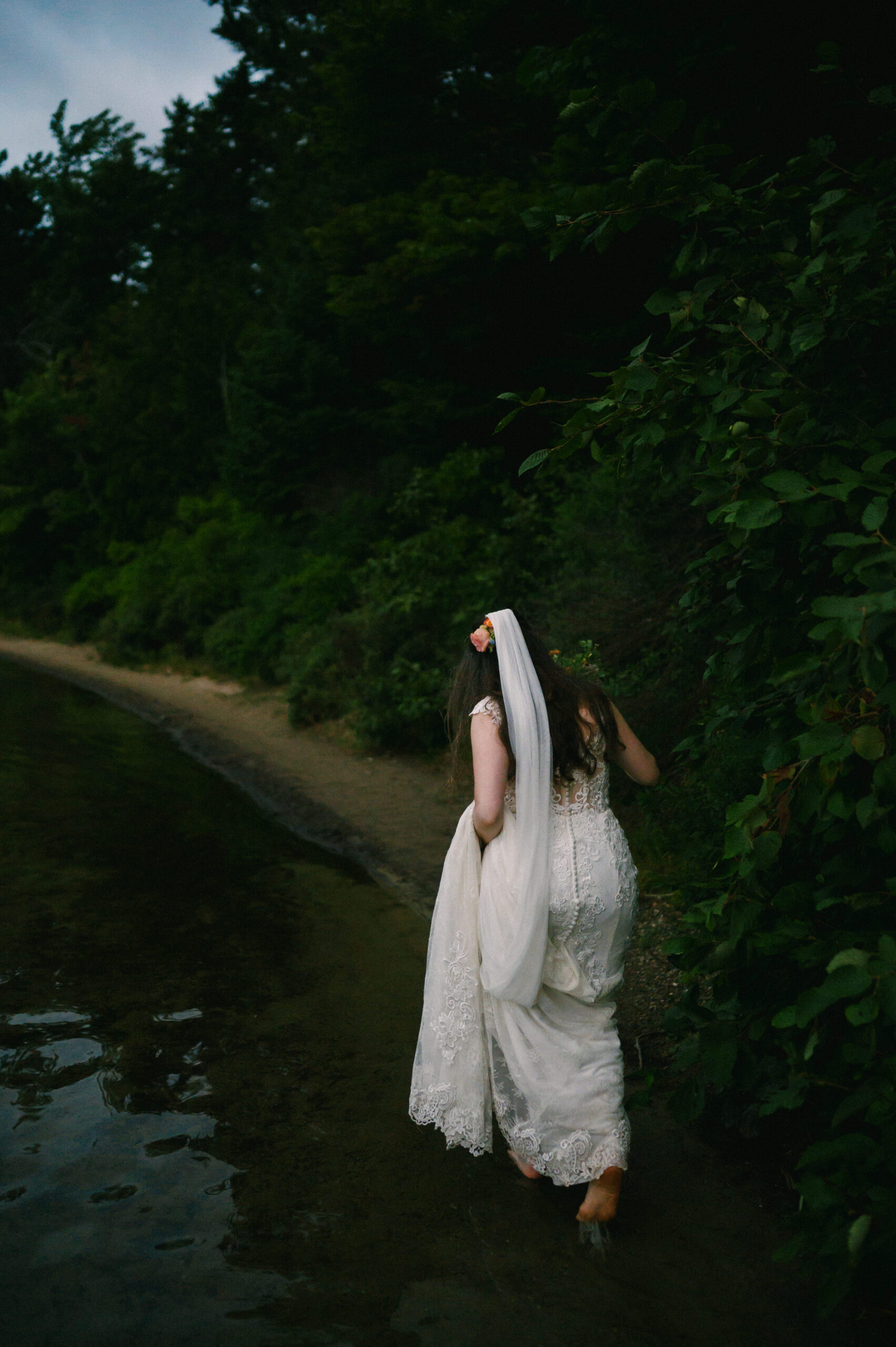 A bride in a lace wedding gown walks barefoot along the edge of a tranquil lake, holding up her dress as she moves along the sandy shoreline, surrounded by dense greenery.