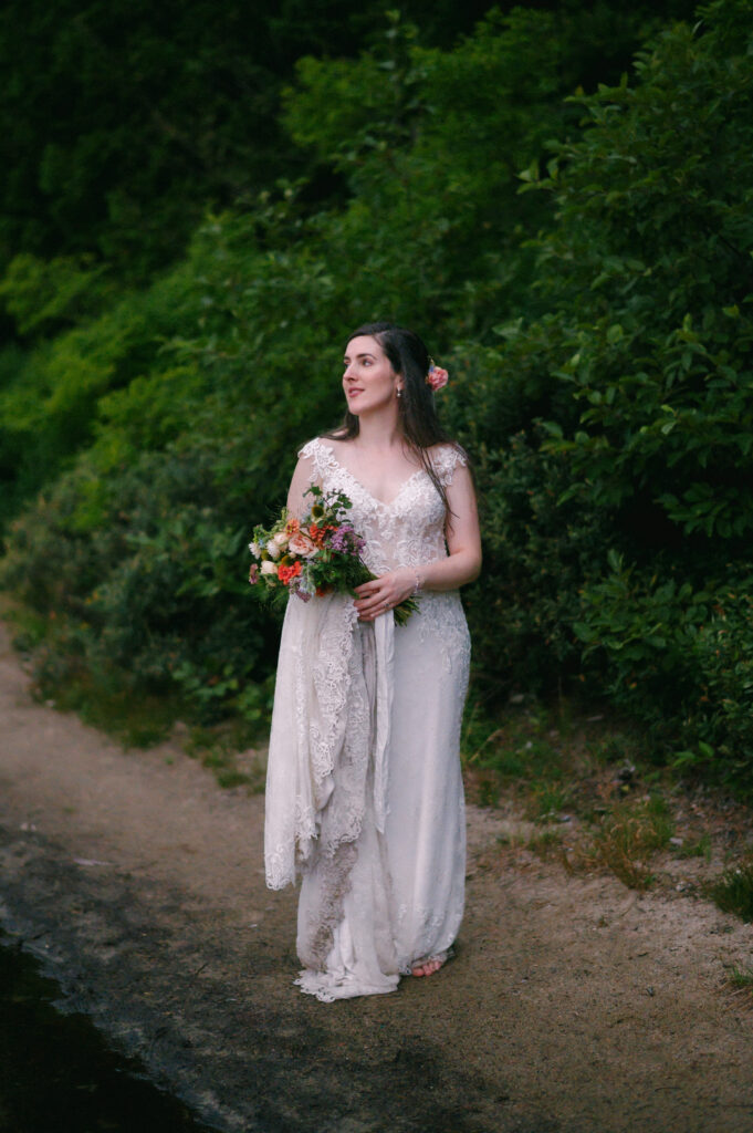 The bride, barefoot and holding her bouquet, walks along the edge of the lake, her lace gown trailing behind her.