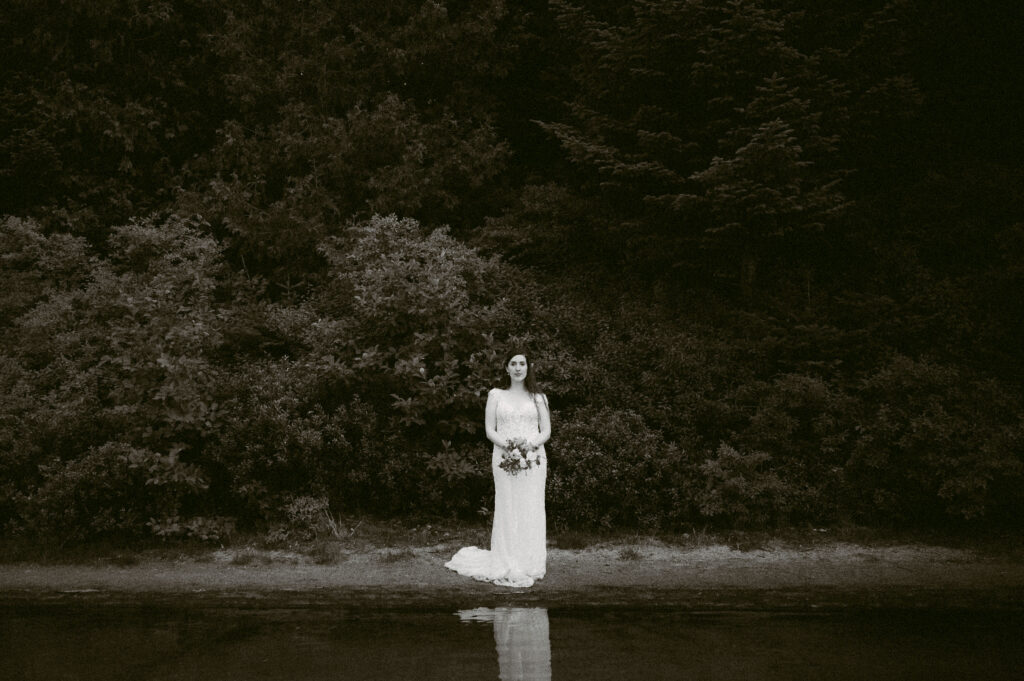 A dramatic black-and-white image of the bride standing alone against a backdrop of dense foliage, her reflection visible in the water.