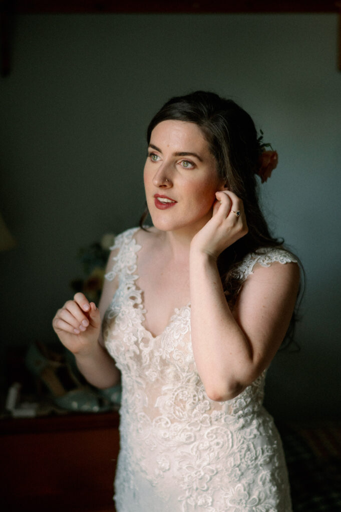 A bride stands in soft, natural light as she adjusts her earring, her expression calm and reflective. She wears a lace wedding gown with delicate floral details, and her dark hair is adorned with a peach-colored flower. The intimate setting of the room, with warm wooden furniture and soft shadows, adds a sense of nostalgia and quiet anticipation.