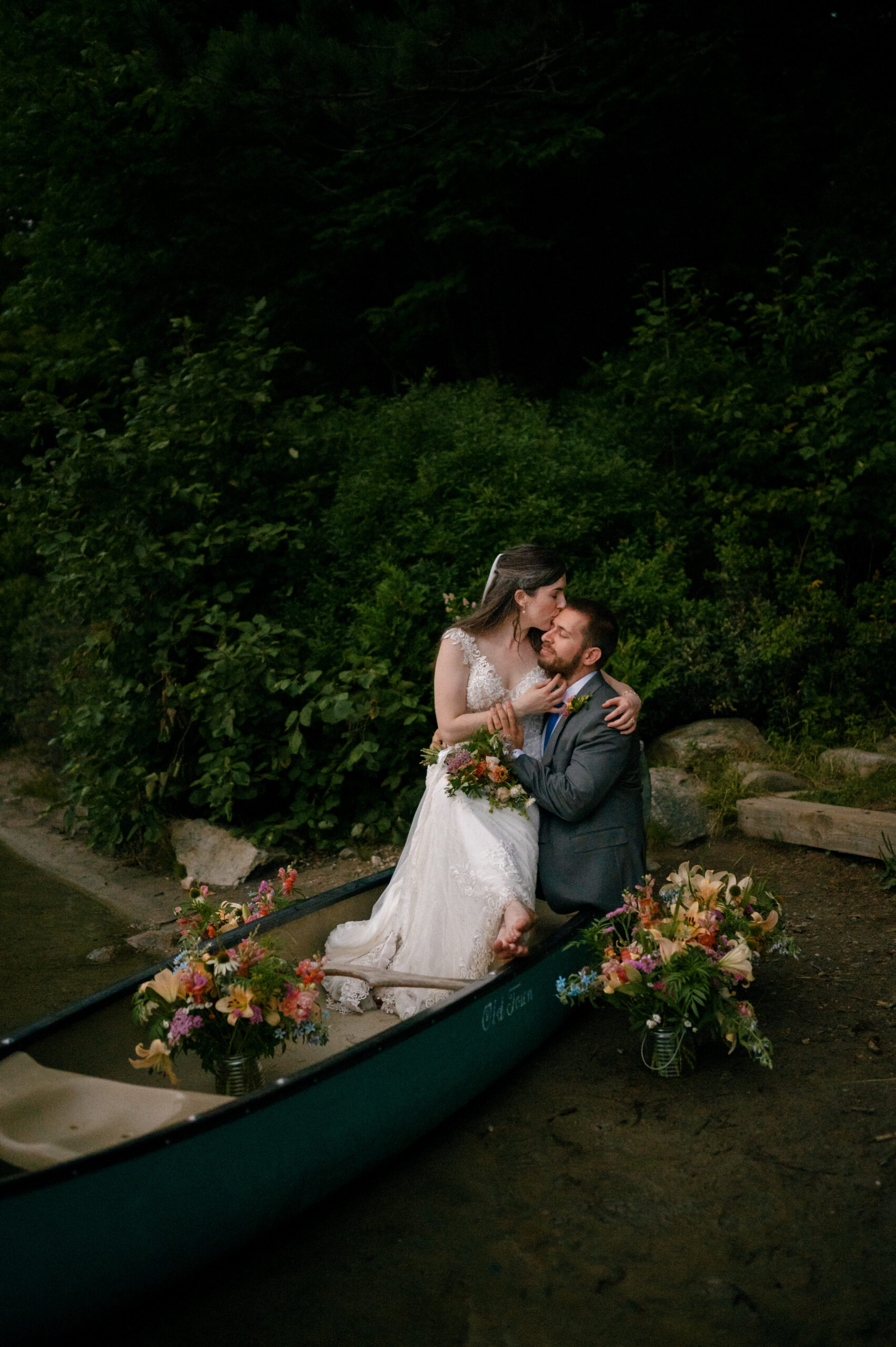 The bride and groom sit in a canoe decorated with colorful flowers, sharing an intimate moment as the bride leans in to kiss her groom’s forehead.