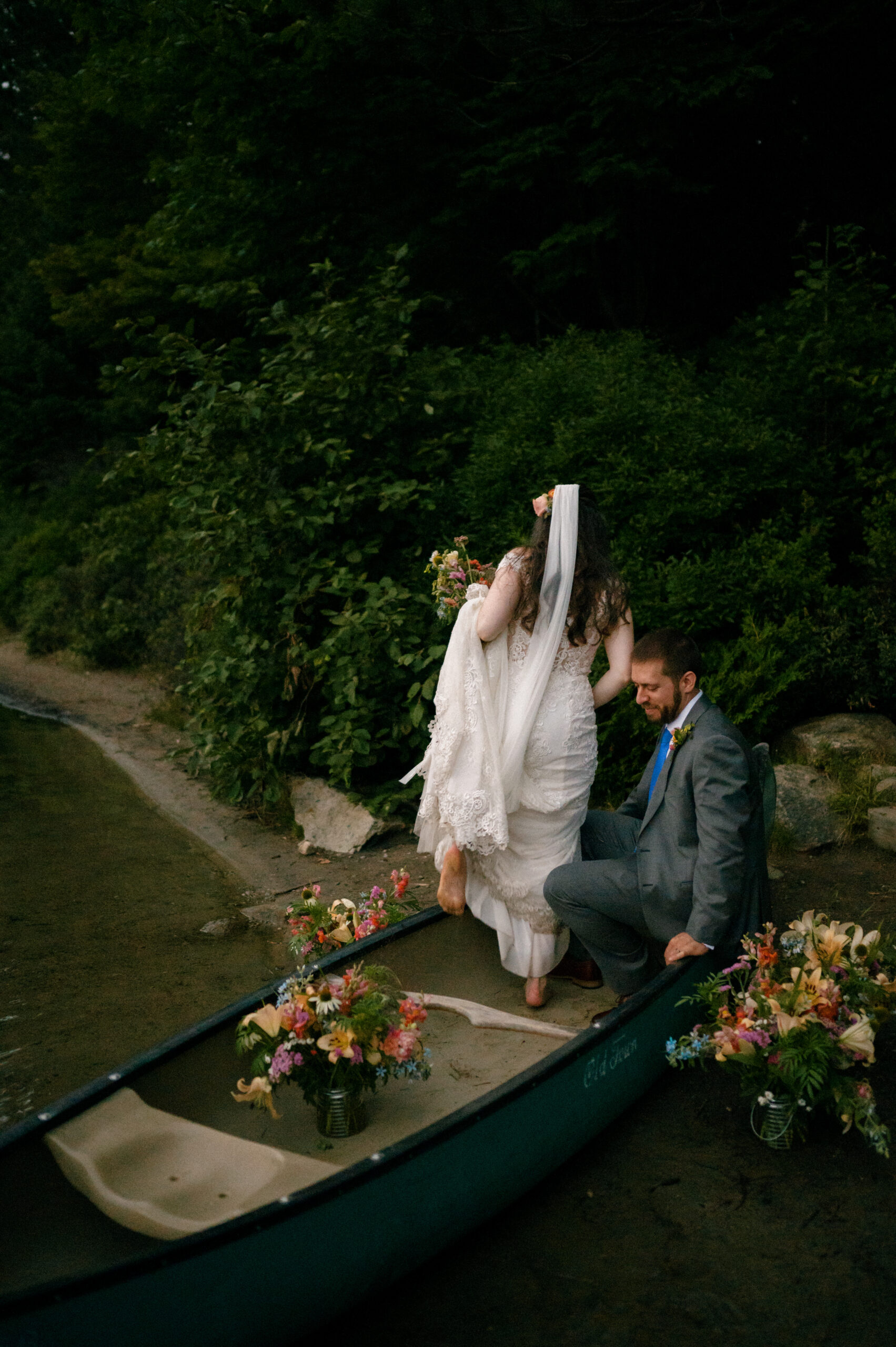 A barefoot bride steps delicately into a canoe adorned with lush floral arrangements, assisted by her groom who sits inside, smiling. Her lace gown trails behind her, and her veil flows gracefully. The couple is surrounded by dense greenery, with the water’s edge visible in the background, creating an adventurous yet romantic scene.