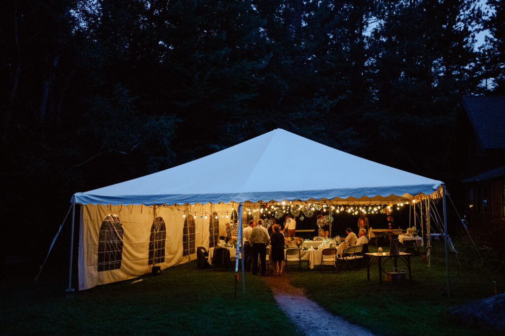 A warmly lit wedding reception under a white tent at twilight, nestled in a wooded setting. String lights cast a golden glow over guests seated at round tables, engaged in conversation and enjoying the intimate atmosphere. The dark blue evening sky and surrounding trees create a cozy, secluded ambiance.