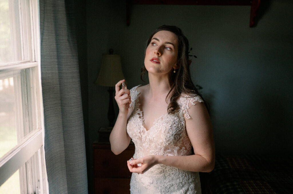 A bride lifts a perfume bottle to spritz her wrist as she stands by a window, bathed in natural light. Her lace gown catches the sunlight, and the nostalgic bedroom setting adds warmth and familiarity to this quiet pre-ceremony moment.