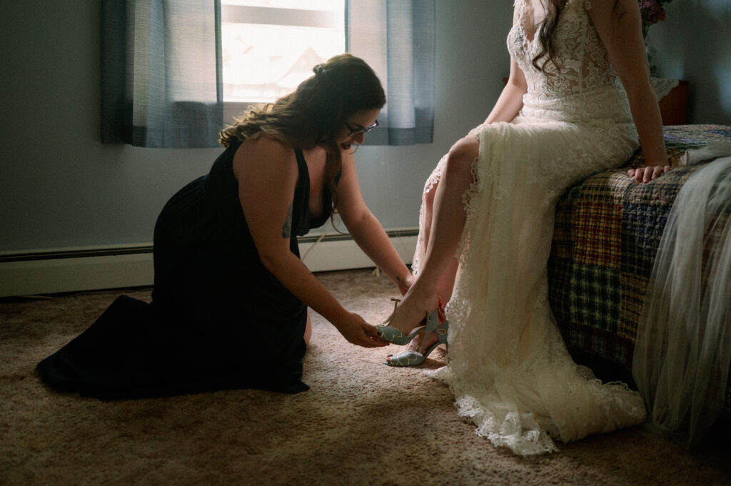 A bridesmaid kneels to help the bride fasten her vintage-inspired teal heels. The bride sits on a quilted bedspread, the soft light filtering through the window creating a warm, nostalgic atmosphere as they share this quiet, meaningful moment before the ceremony.