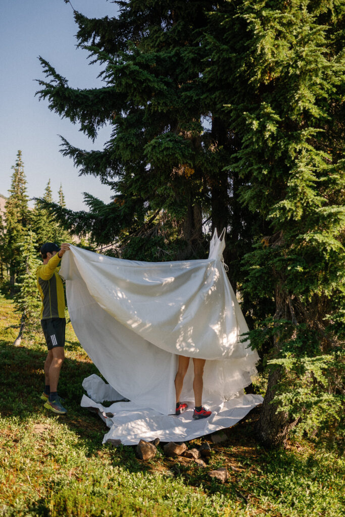 Backcountry elopement moment of the bride changing behind a white dress held up by a friend in the trees of Goat Rocks Wilderness.
