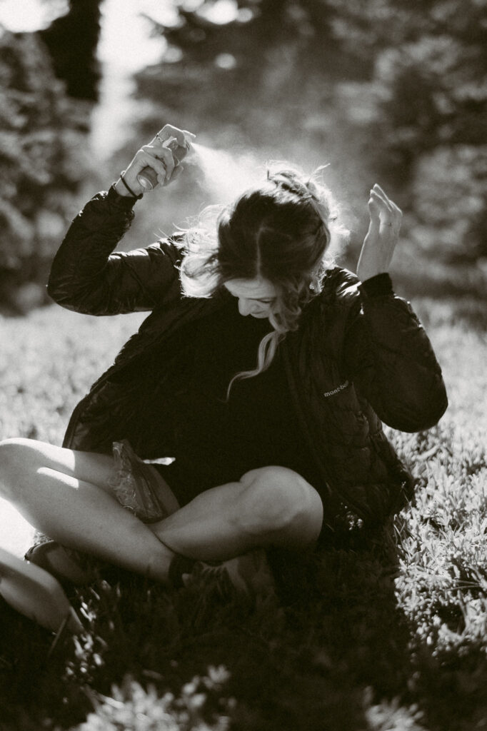 Bride applying hairspray while sitting in the grass, getting ready for her backcountry wedding in Goat Rocks Wilderness.