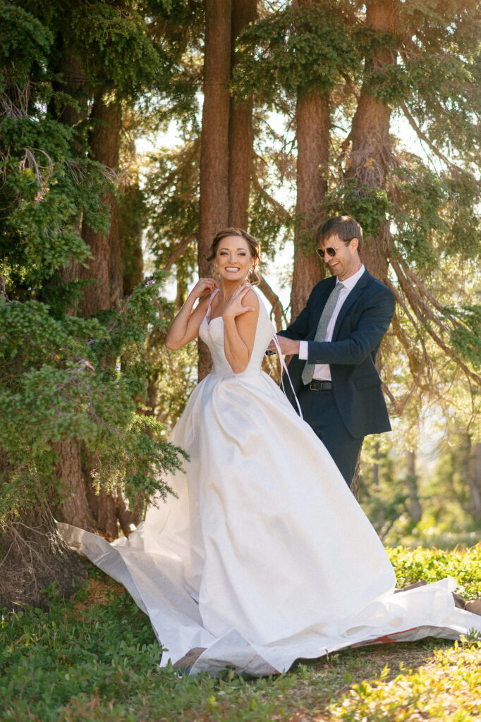 Bride smiling as her friend helps tie the back of her wedding dress during a joyful mountain elopement prep scene.