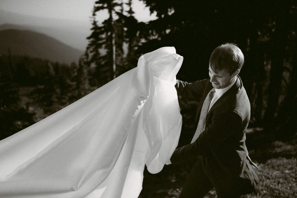 Black and white photo of a friend lifting bride’s dress to help it flow before her intimate wilderness ceremony in Washington.