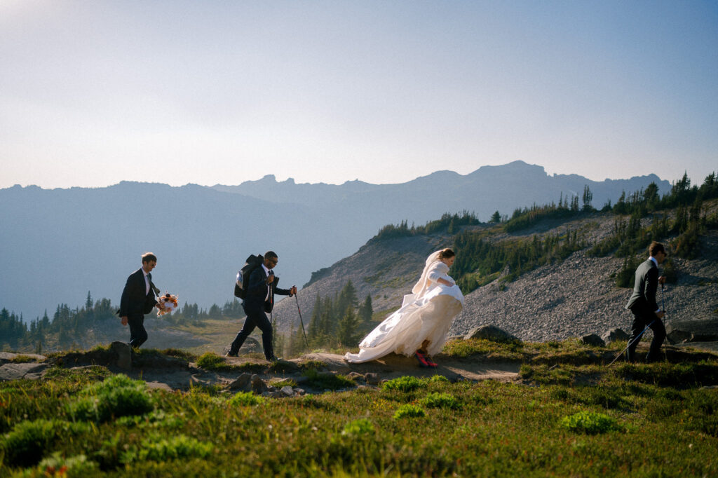 The wedding party ascends the PCT trail—each person in formalwear and hiking gear—backdropped by jagged mountain silhouettes and pine trees.