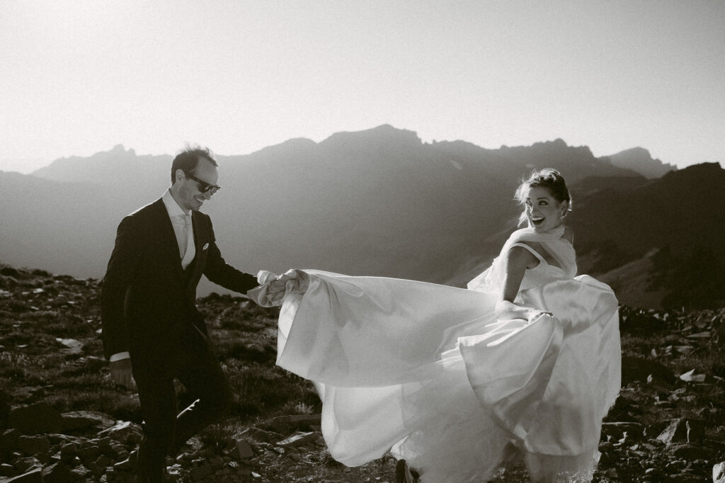 The groom holds the bride’s gown as she looks over her shoulder and laughs, backlit by the fading sun and dramatic peaks in the distance.