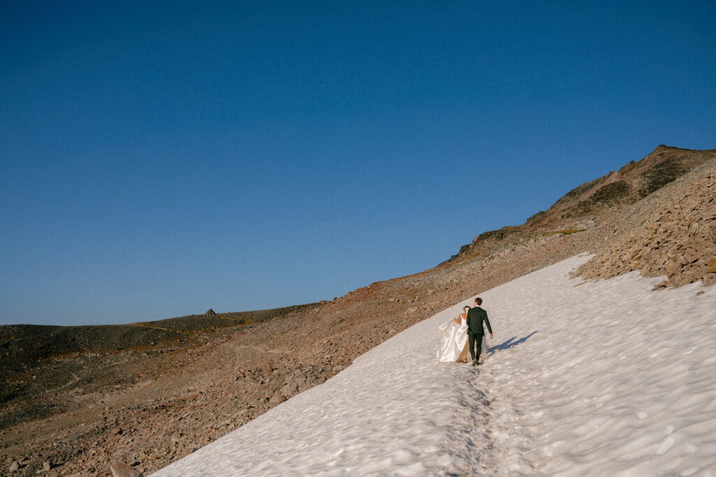 A bride and groom trek hand-in-hand across a snowfield, her white gown trailing behind as they walk toward the rocky slope under a clear sky