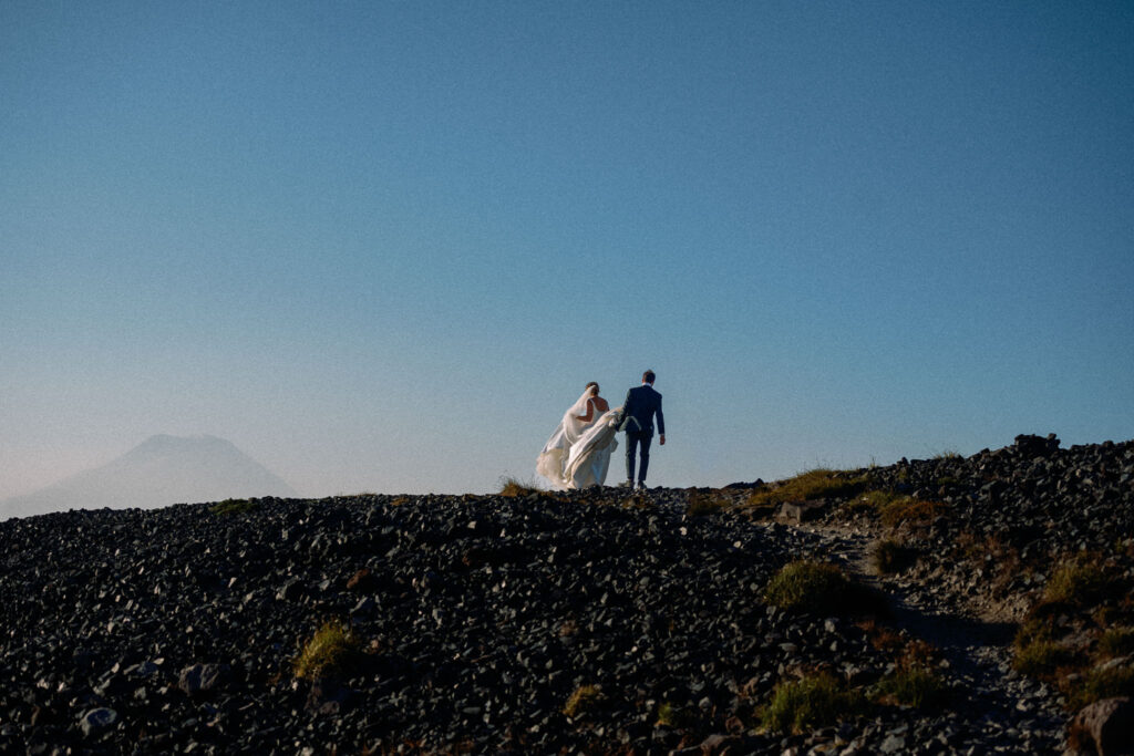 The couple climbs a ridge of dark volcanic rock, silhouetted against a pastel sky with Mount Rainier’s shape barely visible in the mist.