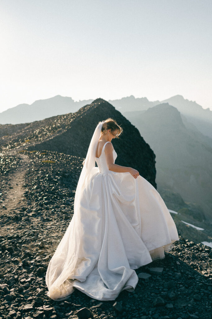 The bride stands alone on the rocky ridge, her dress flowing as she looks down, with layers of jagged mountains fading into soft evening light.