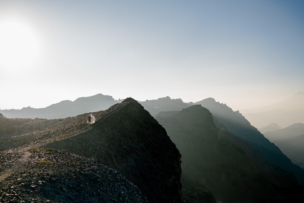 A cinematic scene of the couple standing on a narrow alpine ridge, glowing in sunlight as rugged peaks and shadowed valleys stretch into the distance.