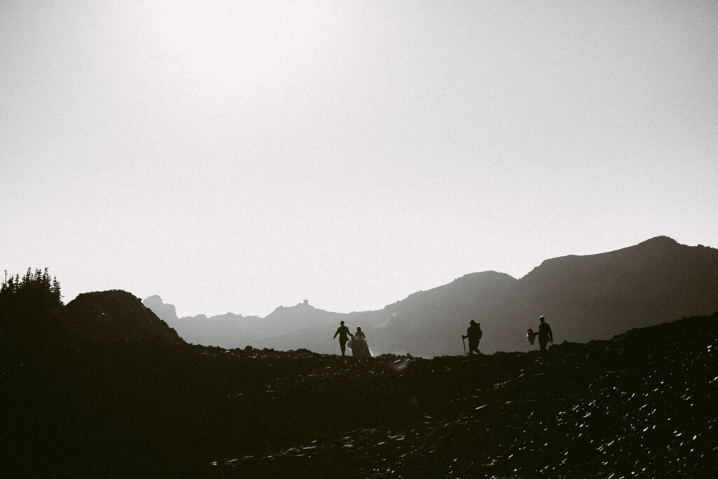 A silhouetted hiking wedding party, including the bride and groom, makes their way along a rocky ridgeline in the Goat Rocks Wilderness at sunrise.