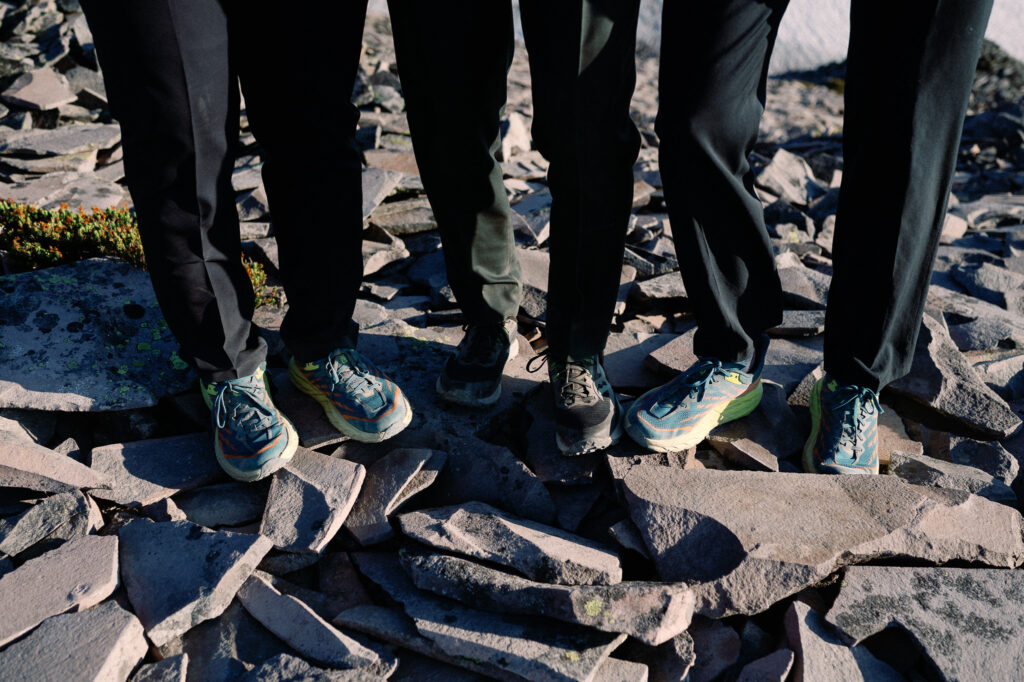 Close-up of four groomsmen’s feet in trail runners and suits, standing on jagged mountain rocks, capturing the blend of formal wear and rugged terrain.