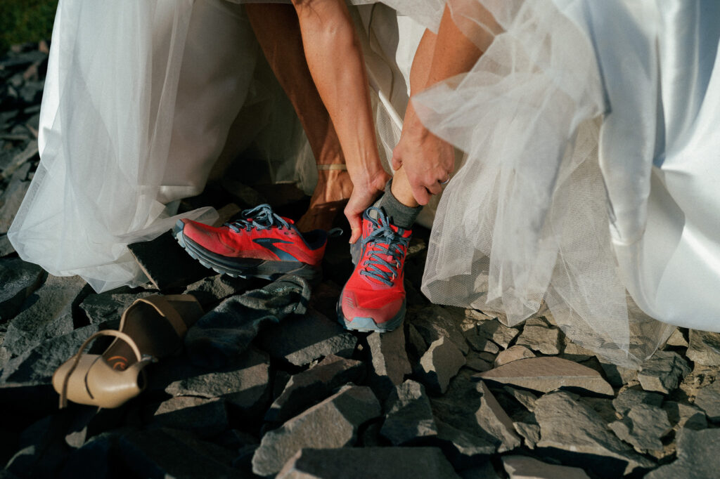 The bride in a tulle wedding dress puts on bright red trail running shoes before continuing the hike, surrounded by loose mountain rocks.