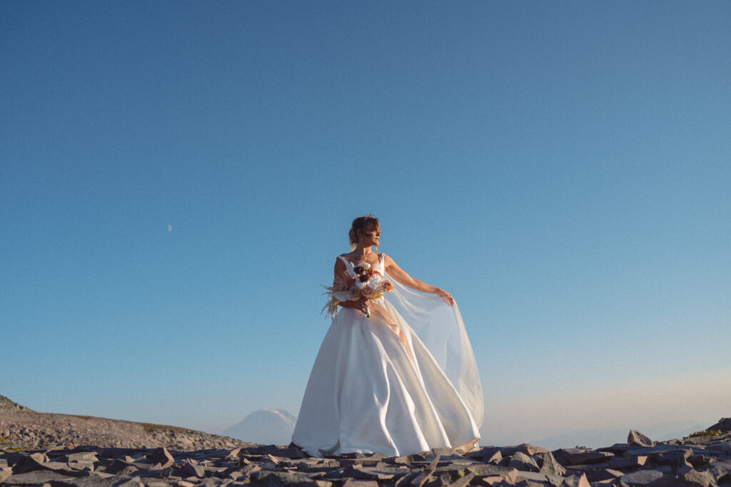 A bride in a flowing satin dress and holding a warm-toned bouquet poses against a vivid blue sky with Mt. Rainier visible in the distance.