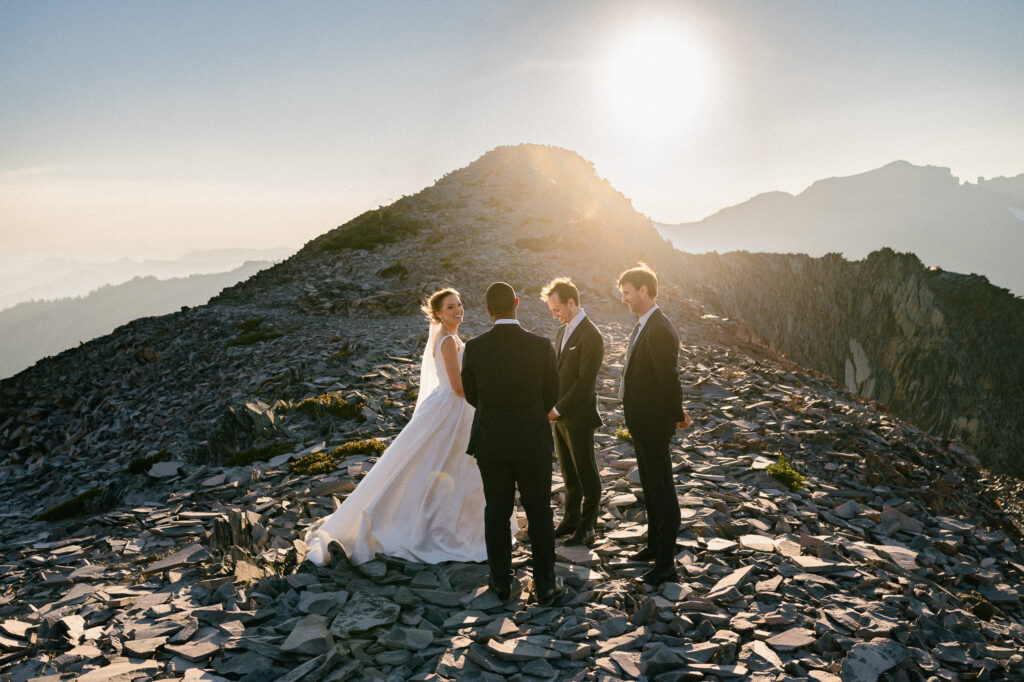 The bride and groom stand with their officiant and best man on a rocky mountain ridge at golden hour, preparing for their mountaintop ceremony.