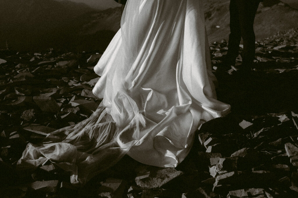 A black-and-white image of the train of the bride’s satin and tulle dress flowing over a bed of jagged mountain stones.