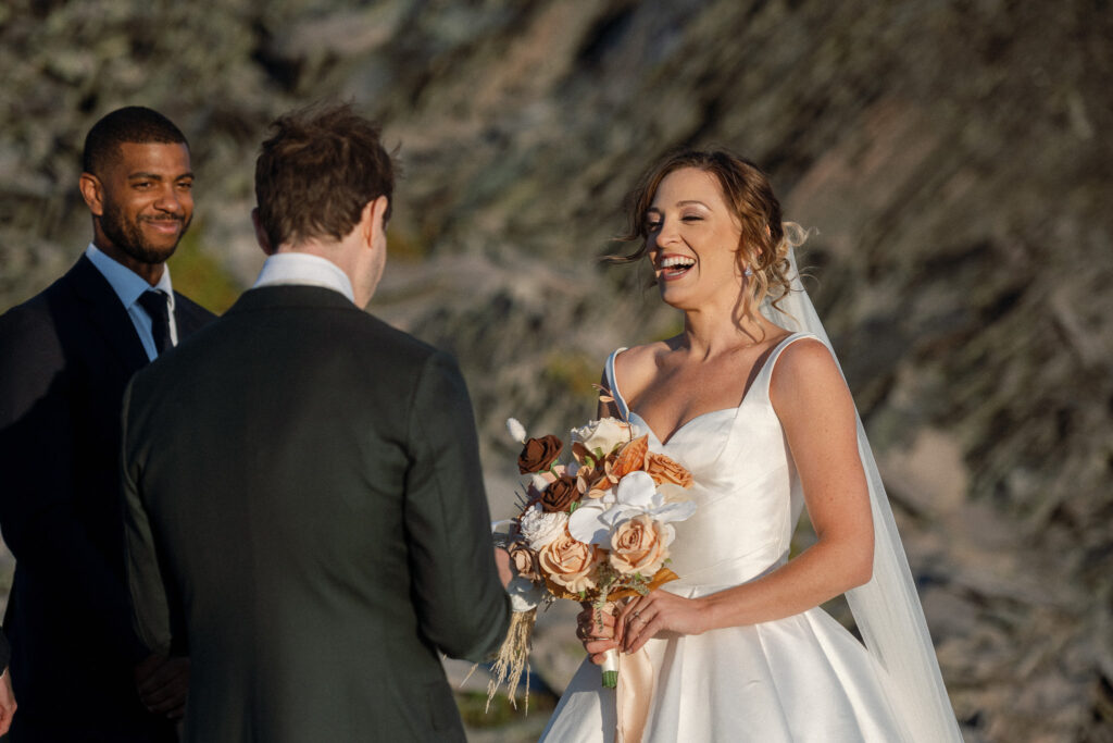 Candid moment during the ceremony with the bride laughing, bouquet in hand, as her groom says his vows with the officiant smiling in the background.