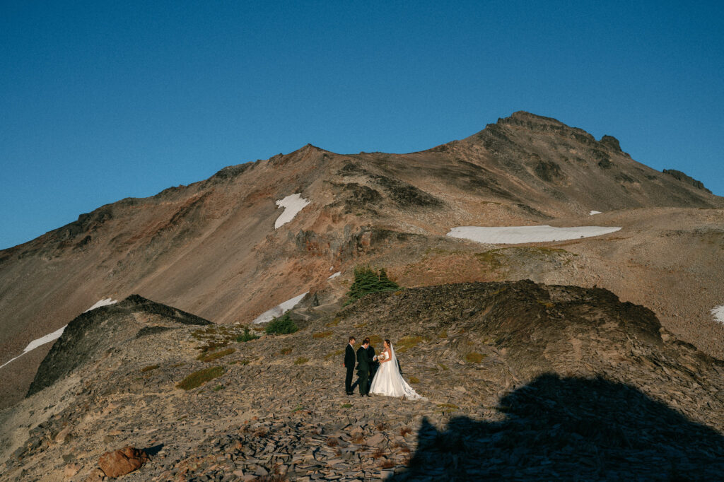 Wide-angle view of a mountaintop elopement ceremony in front of rugged Goat Rocks peaks and patches of lingering summer snow under a clear sky.