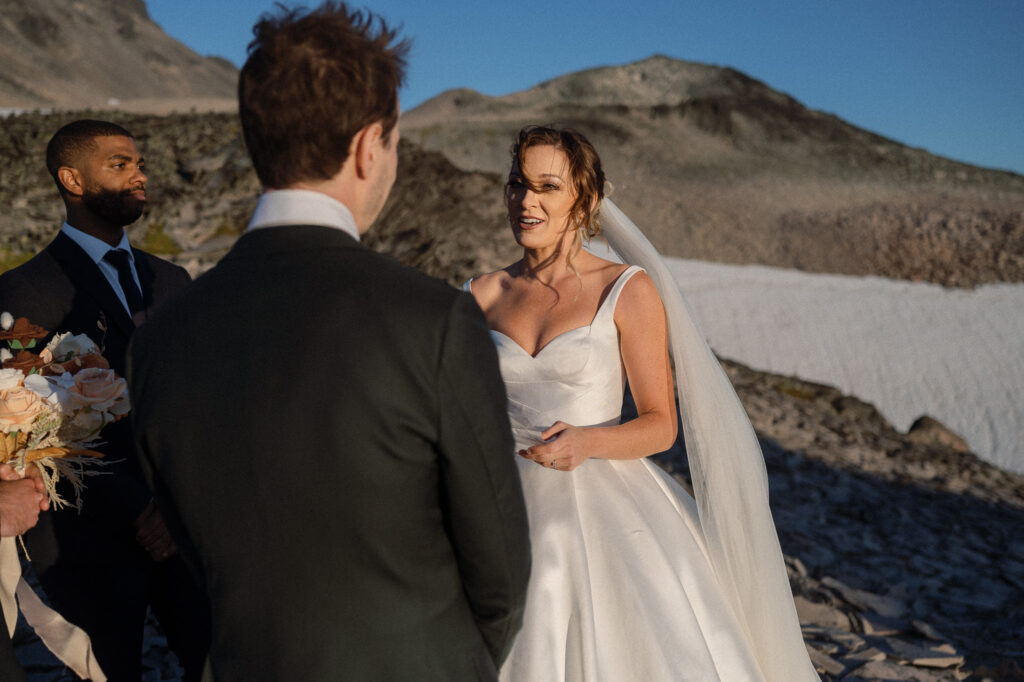 The bride speaks her vows with emotion and confidence as the groom listens, surrounded by rugged alpine terrain and patches of snow.