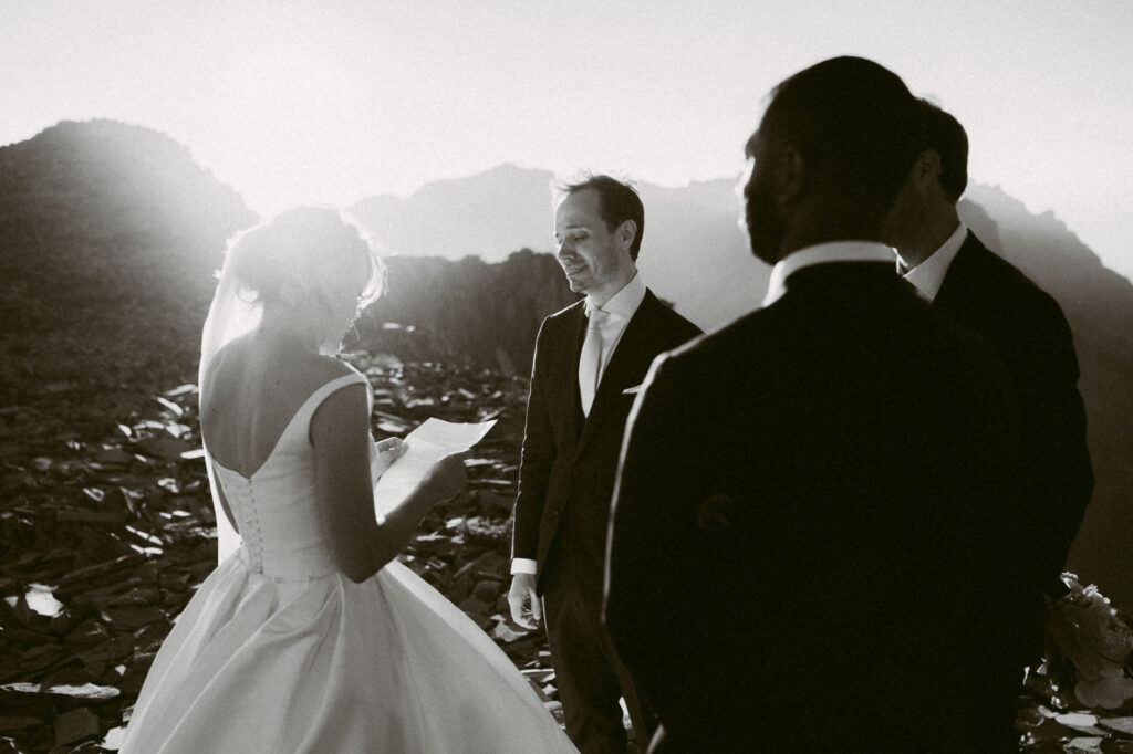 The bride reads her vows to the groom during an emotional mountain-top ceremony, golden sunlight streaming in from behind her