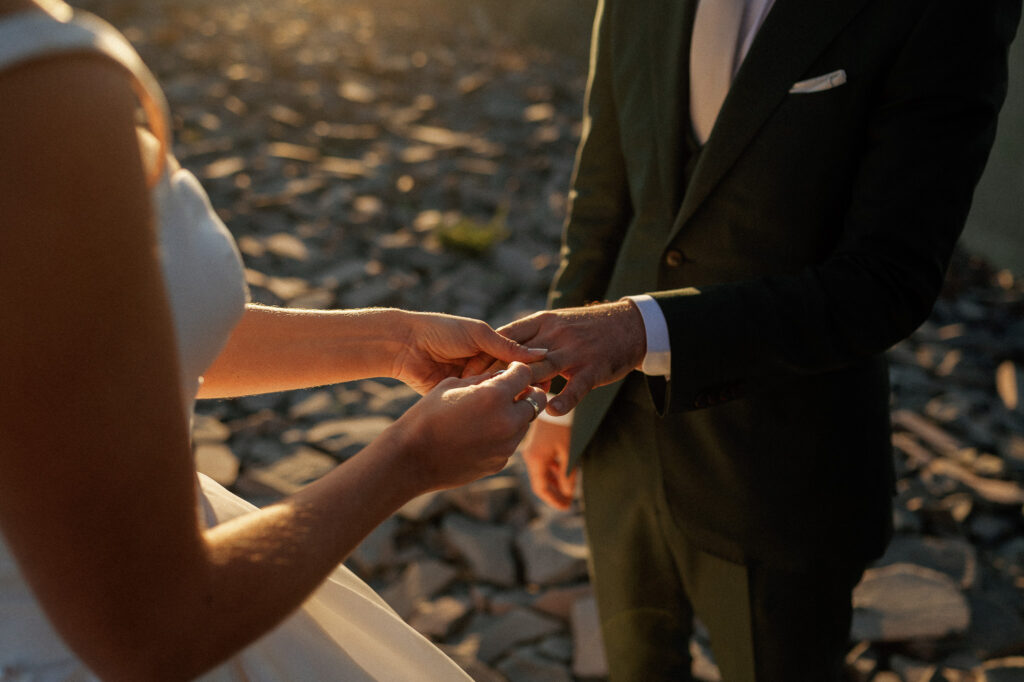 A close-up of the bride slipping the wedding ring onto the groom’s finger, sunlight casting a warm glow on their hands.