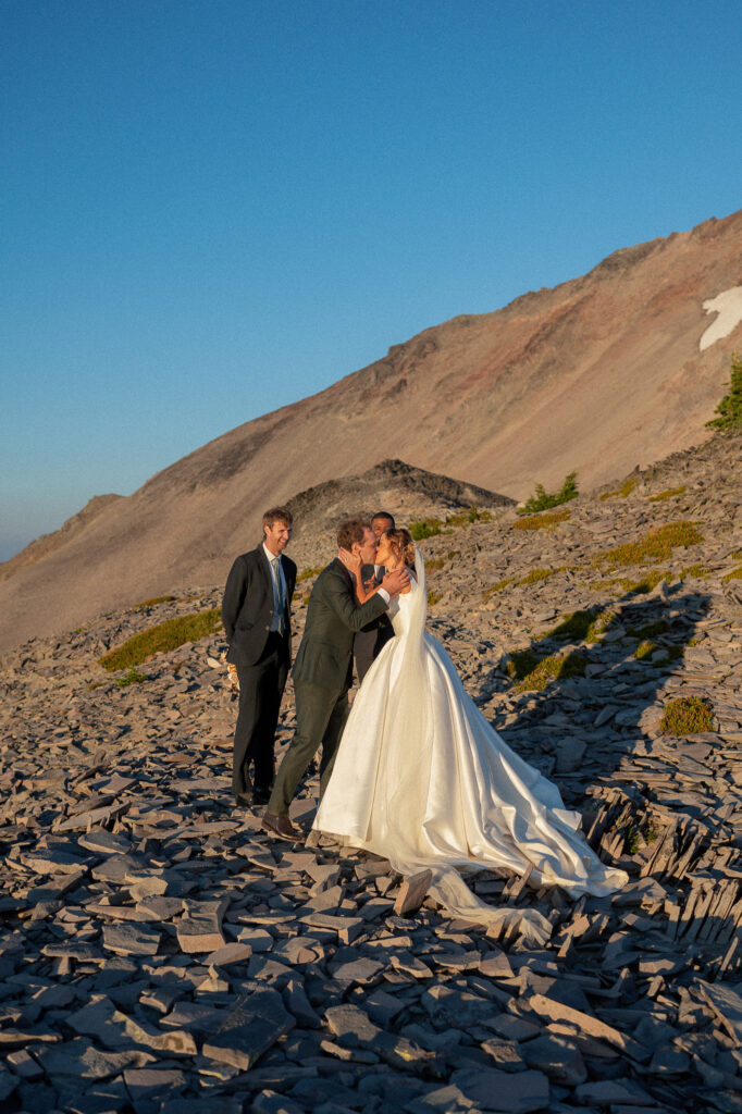 The couple shares their first kiss as newlyweds on a rocky ridge, with their officiant and witness smiling nearby beneath a bright blue sky.