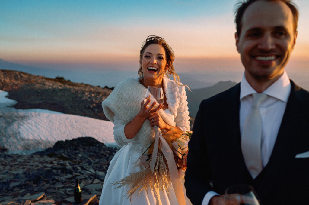Bride laughs warmly while holding a bouquet and wrapped in a white shawl, enjoying a toast after the ceremony.