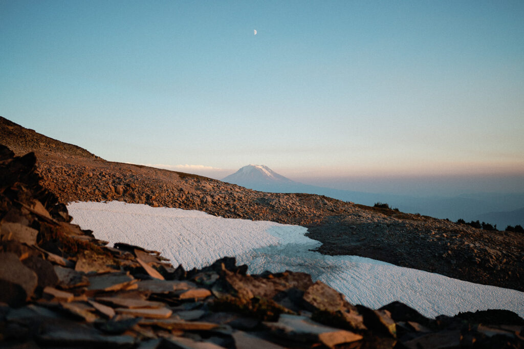 A scenic view of Mount Adams in the distance, with glacier patches in the foreground under a pastel evening sky.