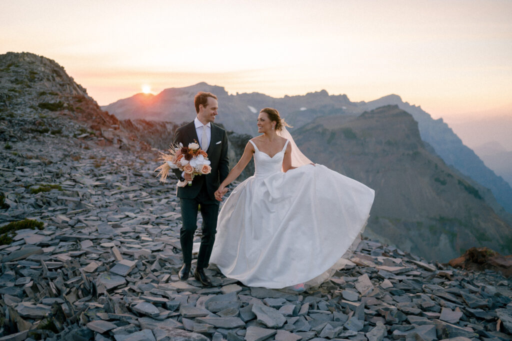 Bride and groom walk hand-in-hand across a rocky ridgeline at sunset, her dress billowing as the sun dips behind the mountains.