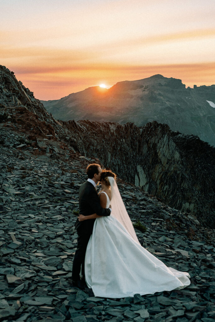 Wide shot of the couple kissing as the sun dips below the rugged mountain peaks in the background.