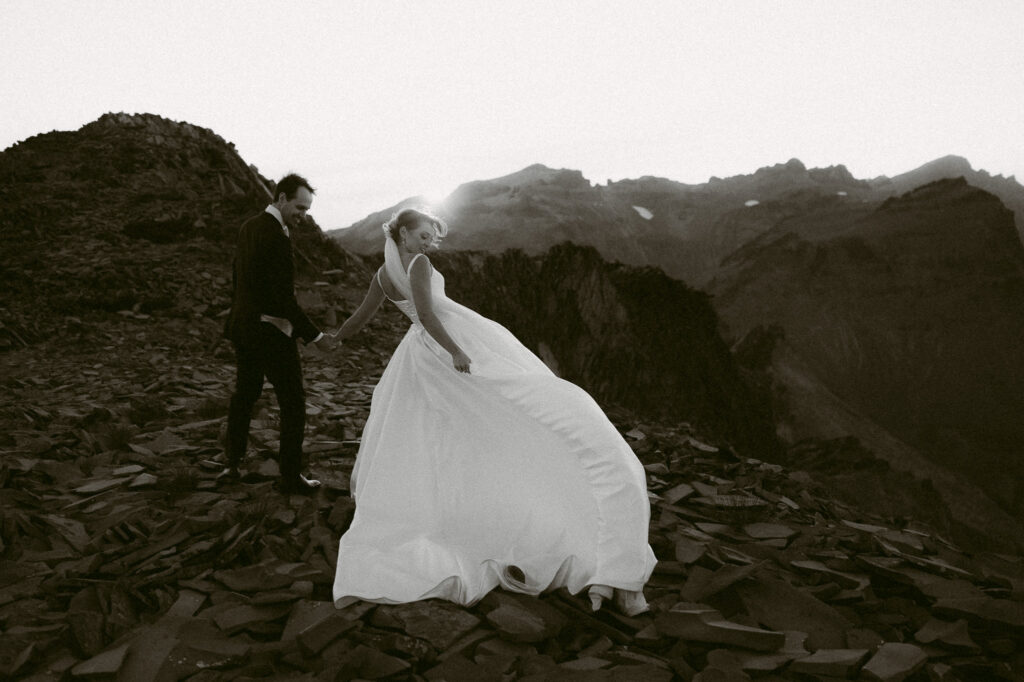 Bride looks back joyfully at the groom as they walk along a mountain ridge, her gown catching the breeze.