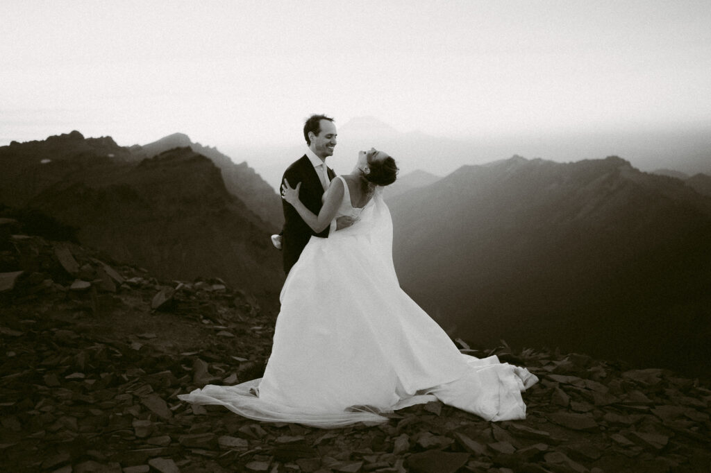 Groom dips the bride as she laughs, silhouetted by distant mountain peaks in the fading light.