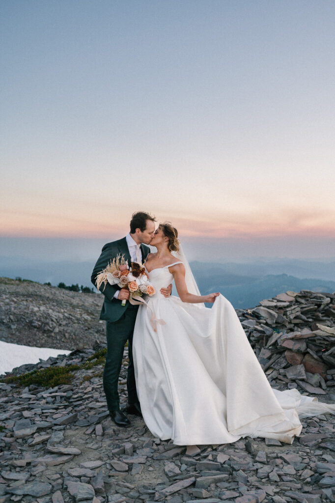 Bride and groom kiss at sunset on a mountain ridge, the bride holding up her gown with one hand and bouquet in the other.