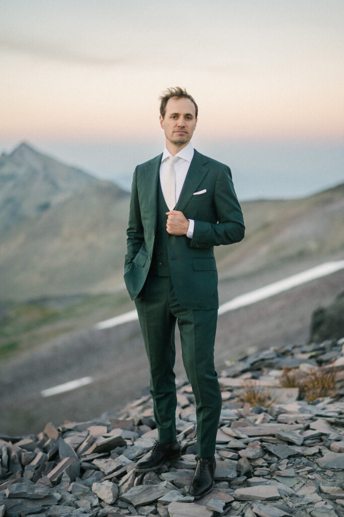 Groom standing alone on rocky terrain at dusk in a dark green suit with hands in pockets.