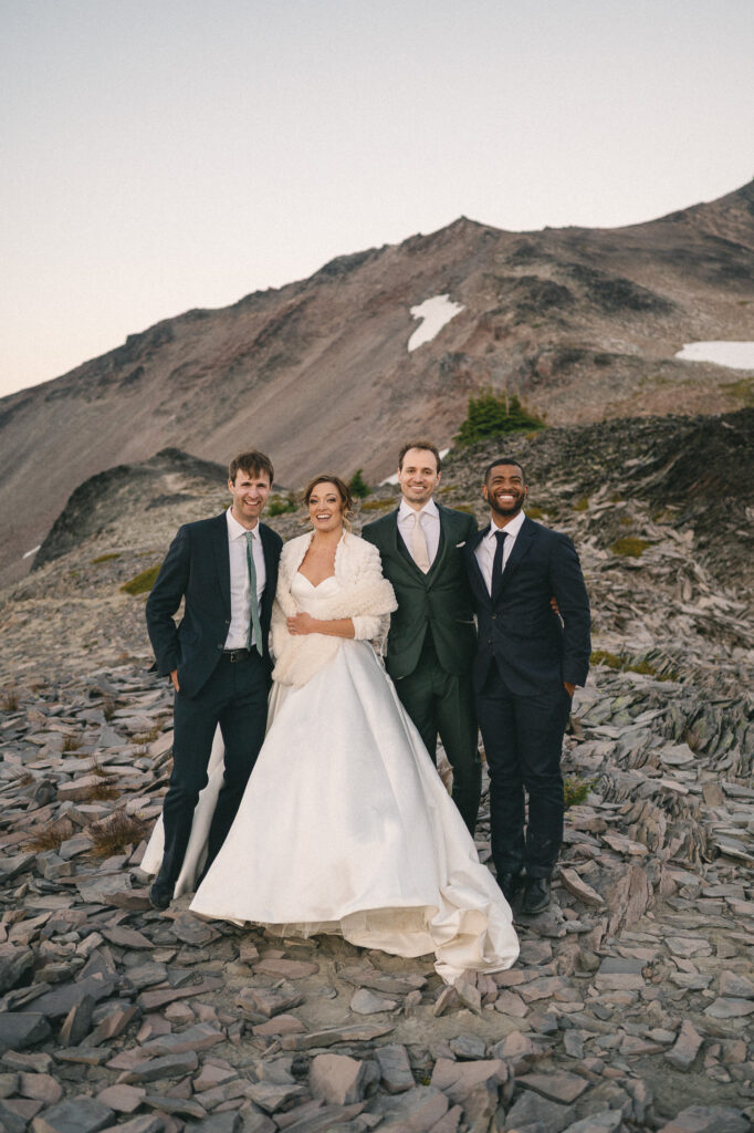Bride and groom pose with two groomsmen on rocky alpine terrain, all smiling and dressed formally.