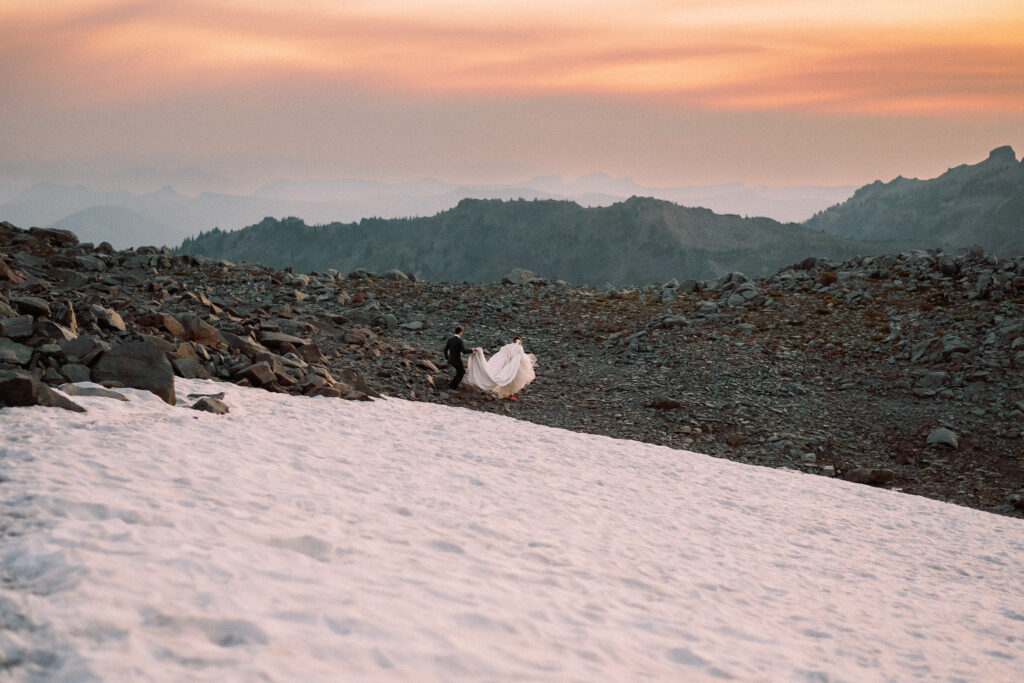Couple walking across alpine terrain near a snowfield, bride holding up her gown with groom assisting.