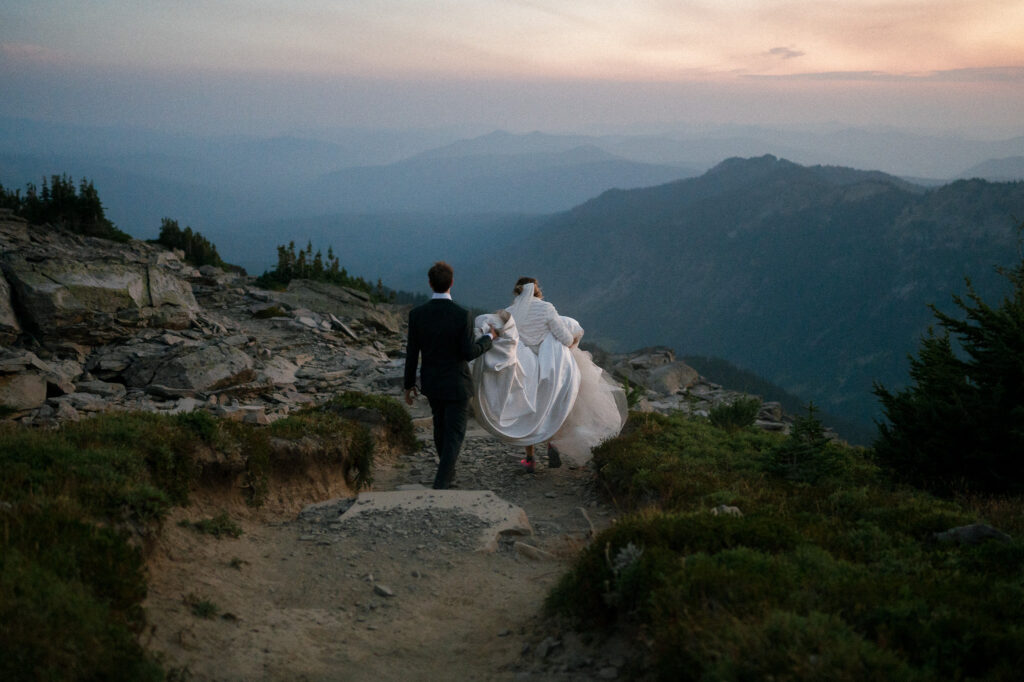 Couple walking down a rugged mountain trail at dusk, bride’s gown trailing as she holds it up.