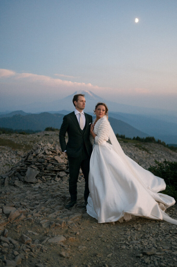 Bride and groom standing still beneath a half-moon sky, the bride’s veil trailing in the wind.