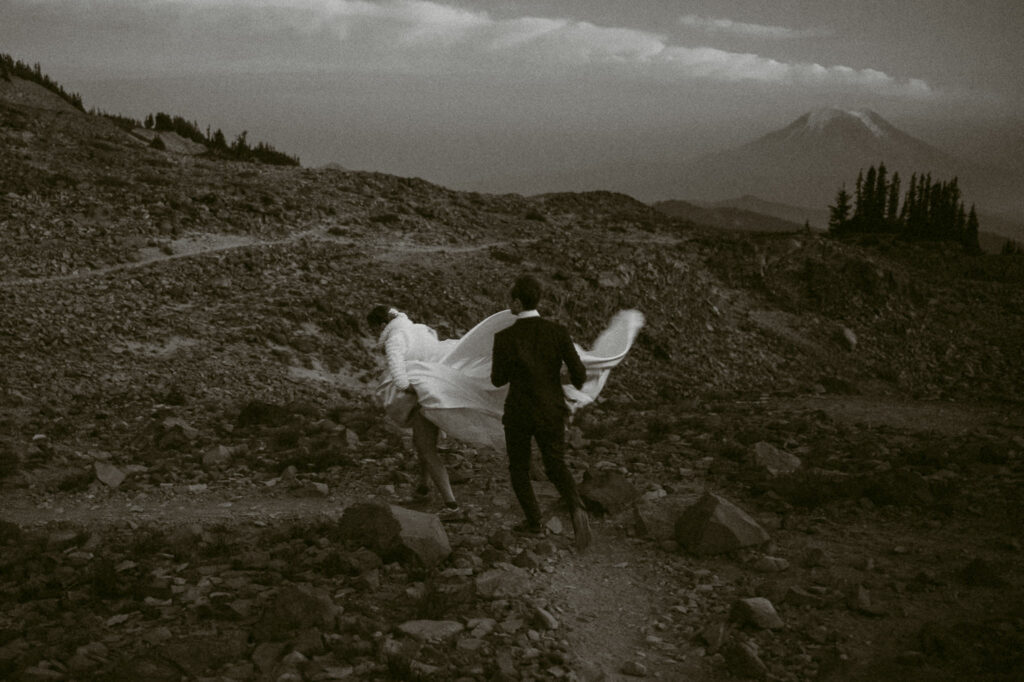 Couple walking down a rugged mountain trail at dusk, bride’s gown trailing as she holds it up.