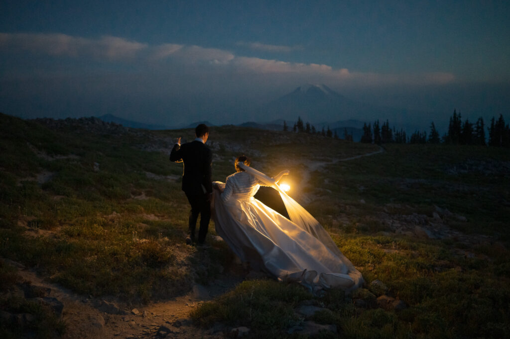 Couple trekking a mountain path by lantern light, bride’s dress and veil illuminated against the twilight landscape.