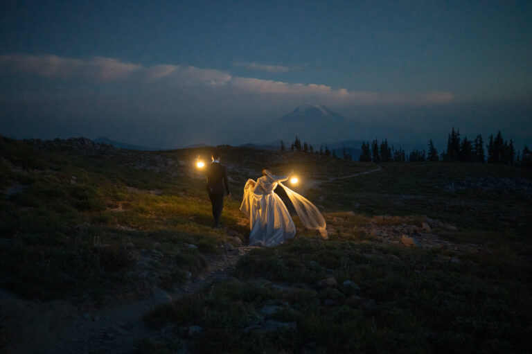 Bride and groom hiking into the night with lanterns glowing, Mount Adams visible in the background beneath a dusky sky.