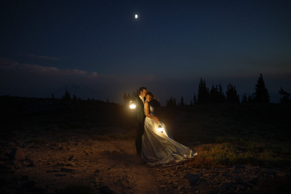 Bride and groom embracing under the moonlight with lanterns in hand, surrounded by silhouettes of distant pines.