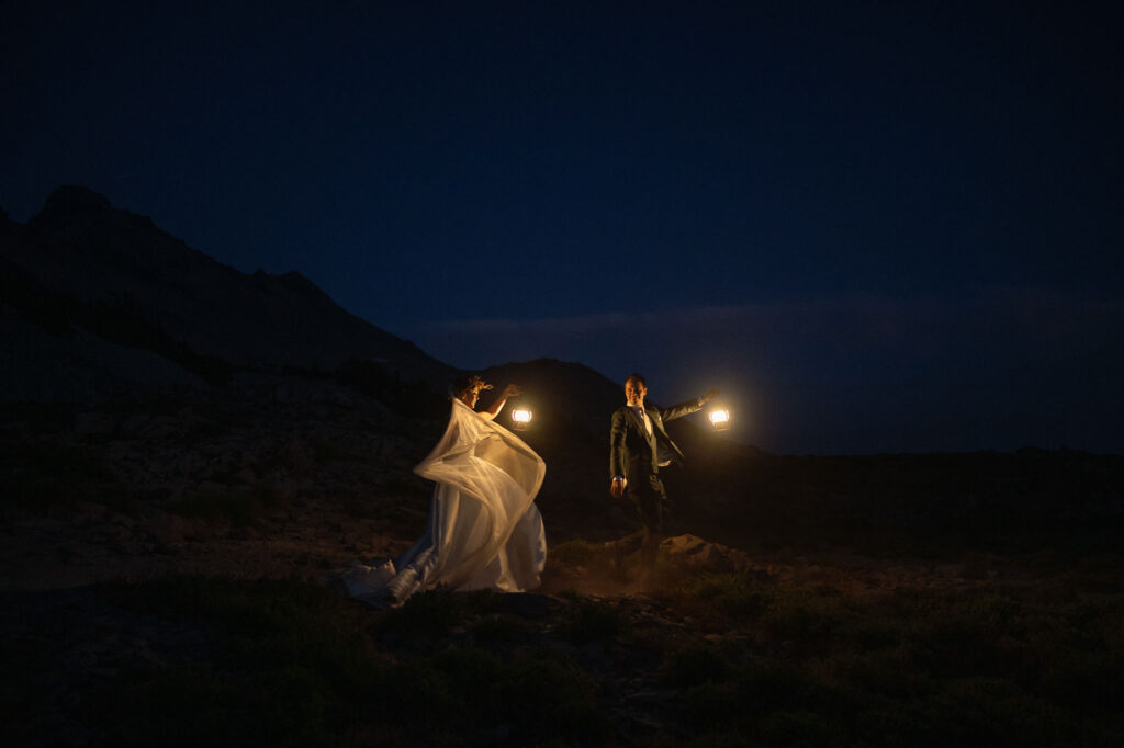 Bride and groom walking hand in hand through the dark wilderness holding lanterns, her veil trailing behind and lit by the warm glow.