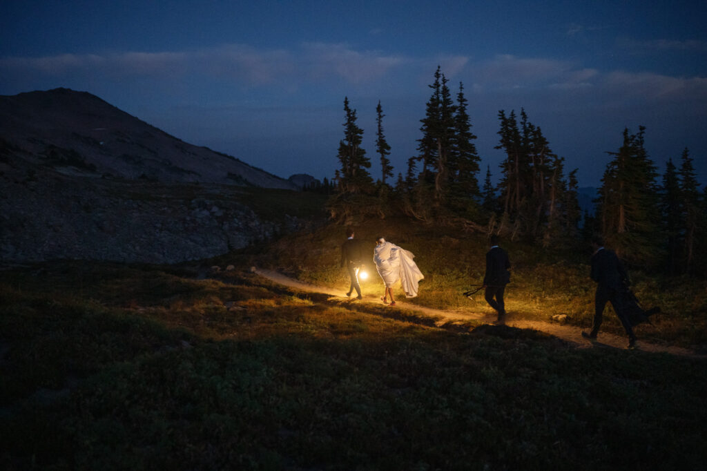Bride and groom silhouetted in front of the trees, each holding a lantern, as they move through the mountain wilderness in the dark.