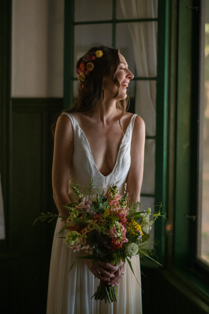 Bride in a deep V-neck gown stands indoors by a window, smiling softly while holding a vibrant, wildflower-style bouquet with colorful blooms and greenery at her summer wedding at Camp Iroquois in the Adirondacks.
